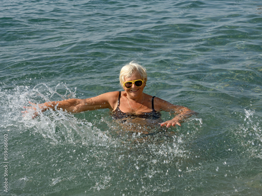Aged woman is doing splashing motions in clear sea water.