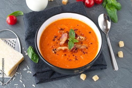 Thick tomato soup with basil and fried bacon in a black ceramic bowl on a grey abstract background. Healthy eating concept Fresh tomato soup with creme fraiche