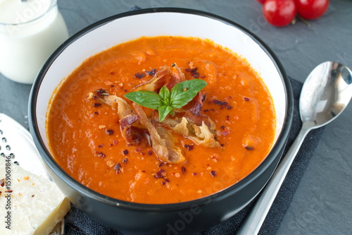 Thick tomato soup with basil and fried bacon in a black ceramic bowl on a grey abstract background. Healthy eating concept Fresh tomato soup with creme fraiche