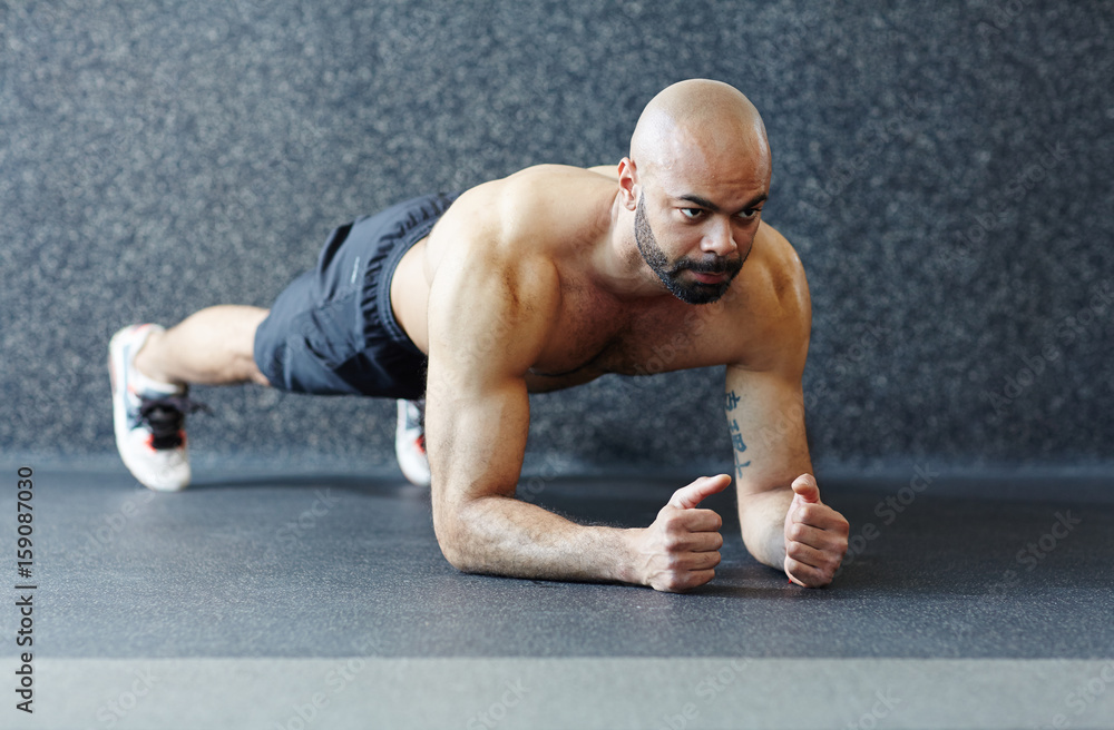 Obraz premium Motivational portrait of shirtless muscular man holding plank against grey background, straining with effort and looking determined during intense endurance workout in gym