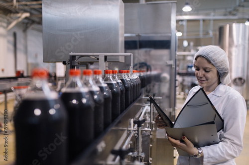 Foto Young happy female worker in bottling factory writing notes about bottles before shipment