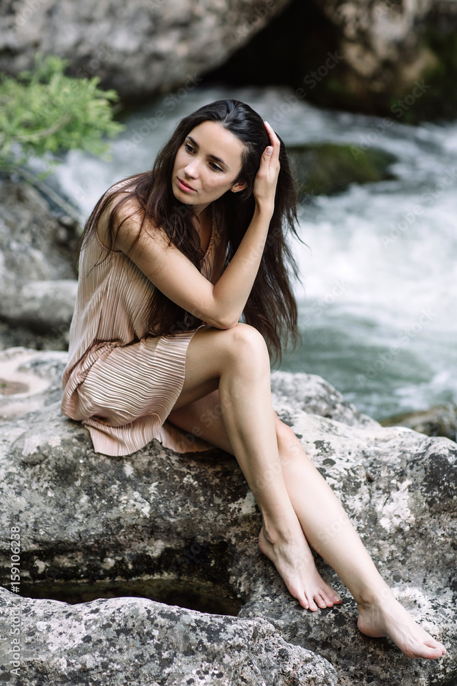 Beautiful brunette sitting on a rock in the middle of the river, water, waterfall. Beautiful nature, natural light, soft, filling