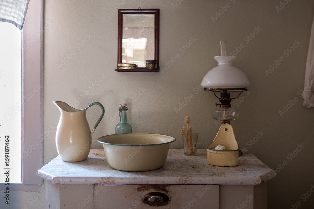 An old wash basin with water jug, wash bowl, soap holder in enamelled  metal. Standing on a commode/chest of drawers with a Kerosene lamp and a  mirror above Stock Photo | Adobe
