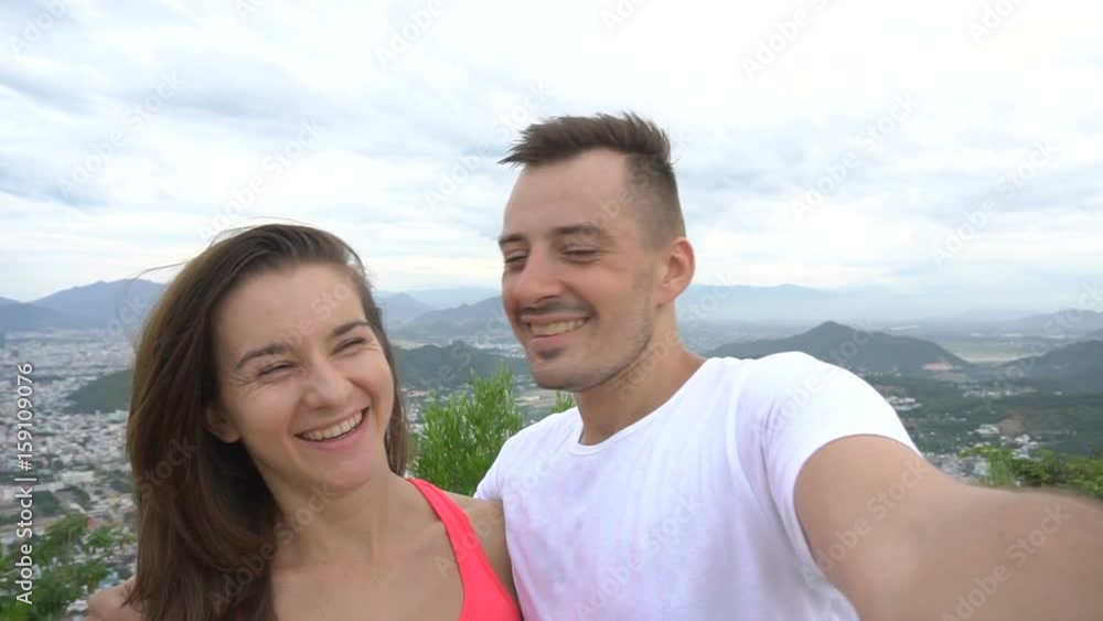 Happy smiling couple recording video, taking selfie in the mountains with aerial city view. Man kisses girl Happy smiling couple recording video, taking selfie in the mountains with aerial city view. Man kisses girl