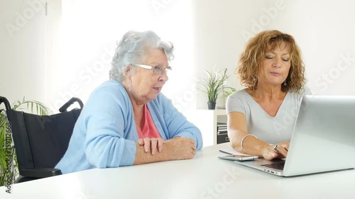 mature woman helping assisted elderly senior female with administrative procedures and paperwork on internet with a laptop computer at home