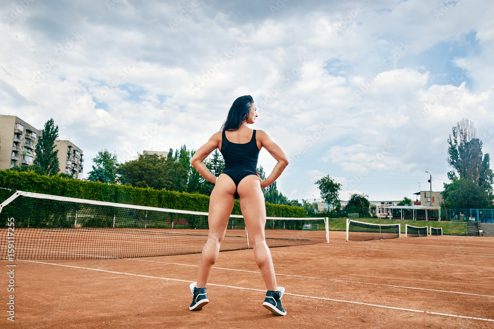 Beautiful young free sports sexy fitness girl dressed in bodysuit, sneakers, with a smart ass and long legs, on a sports ground on a tennis court with a beautiful sky in the background