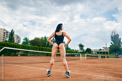 Beautiful young free sports sexy fitness girl dressed in bodysuit, sneakers, with a smart ass and long legs, on a sports ground on a tennis court with a beautiful sky in the background © tavrox