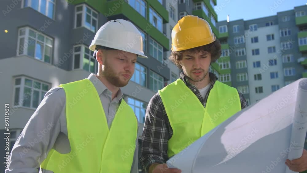 Two caucasian builders in protected helmets and vests looking drawing. Builder standing near his collegue using mobile phone. Outdoor.
