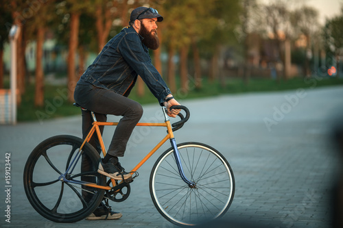 Bearded guy and fix bike. Young man with gourgeous large beard in sunglasses and in the cap riding a vintage bicycle in the city park at sunset.