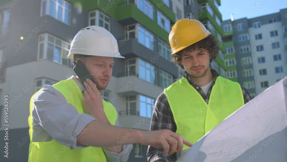 Team of young caucasian builders in protected helmets and vests looking drawing. Builder standing near his collegue using smart phone. Outdoor. Team of young caucasian builders in protected helmets and vests looking drawing. Builder standing near his collegue using smart phone. Outdoor.
