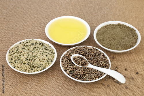 Hemp health food with hulled and dried seed, oil and powder in porcelain bowls with spoon on hessian background. 