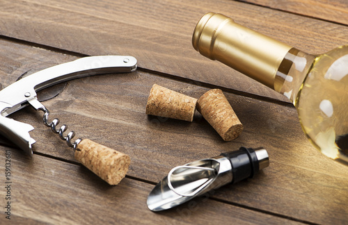Corkscrew next to several corks and a bottle of white wine on wooden table. Horizontal studio shot.