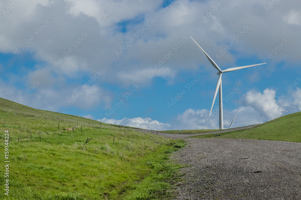 A gravel road leads to a wind turbine on a hillside, against the backdrop of cloud-filled blue sky in the Livermore, California wind farm