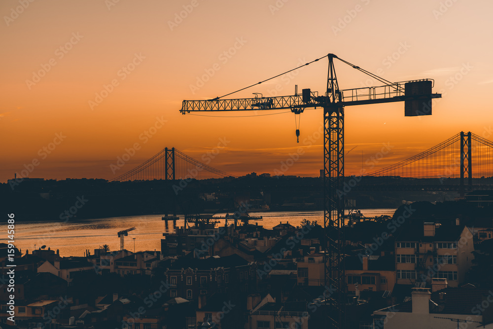 Stunning sunset in Lisbon shooting from high point with silhouettes of construction crane, famous rope bridge, river, multiple facades of old houses in central district and dramatic orange sky Stunning sunset in Lisbon shooting from high point with silhouettes of construction crane, famous rope bridge, river, multiple facades of old houses in central district and dramatic orange sky