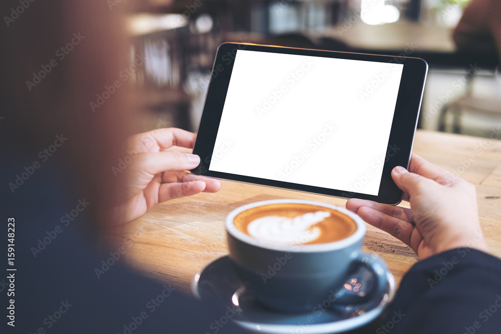 Mock up image of business woman's hands holding black tablet with white blank screen and coffee cup on wooden table in cafe Mock up image of business woman's hands holding black tablet with white blank screen and coffee cup on wooden table in cafe