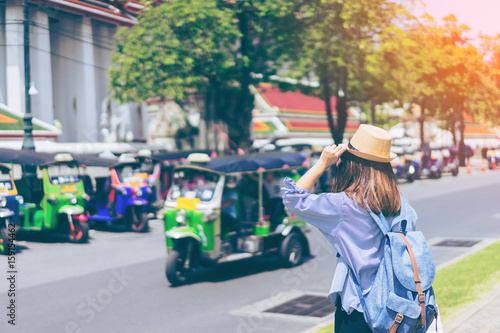 Young woman traveler with sky blue backpack and hat looking the way with tuk tuk thailand background from wat pho at Bangkok Thailand
