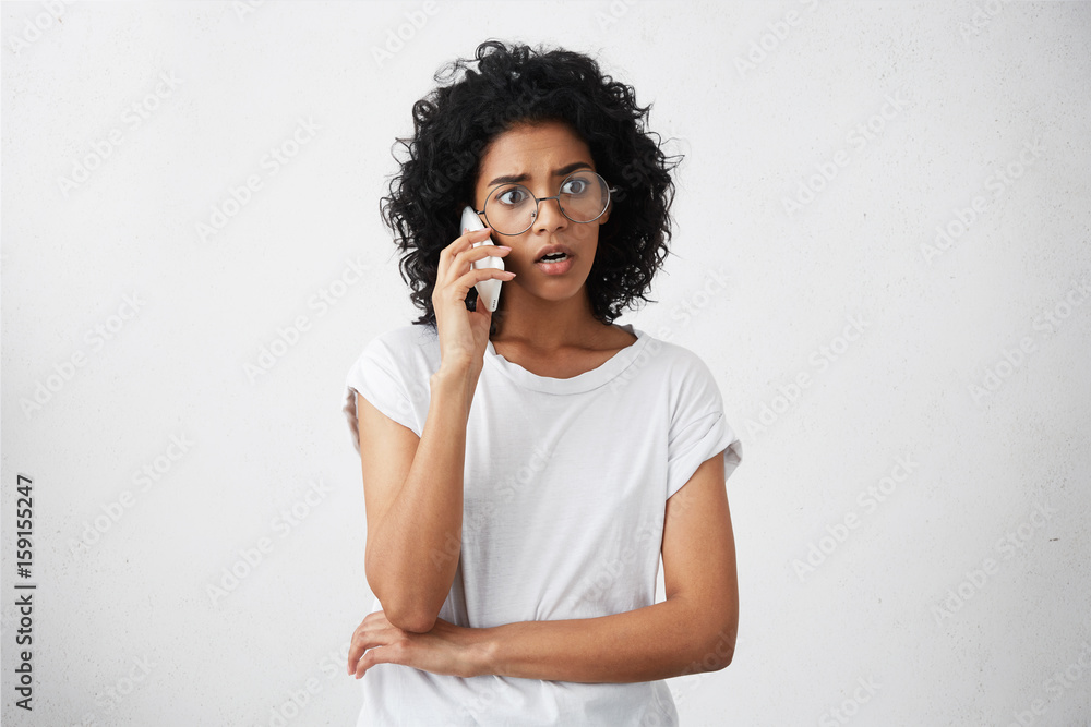 Afro American housewife with curly hair wearing big round glasses and casual T-shirt talking over smartphone with her friend while having free time having surprised look after hearing shocked news Afro American housewife with curly hair wearing big round glasses and casual T-shirt talking over smartphone with her friend while having free time having surprised look after hearing shocked news