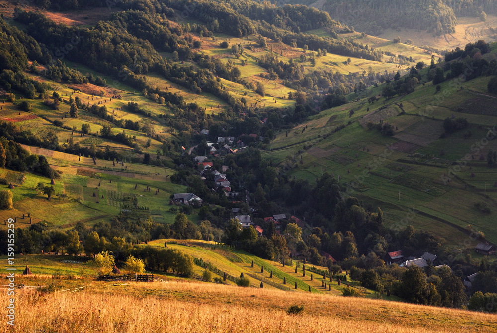 A beautiful summer rural landscape with houses, sunny hills and  many small hay stacks. Carpathian rolling landscape on sunset in autumn colors. Picturesque pastoral Ukrainian landscape. West Ukraine