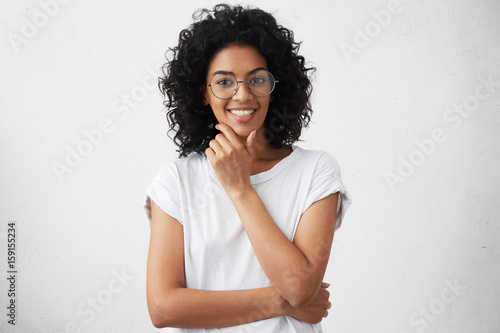 Friendly happy Afriacan American teenager wearing casual T-shirt and big round glasses holding her hand on chin having joyful and cheerful look going to have walk in park with her best friend © wayhome.studio 