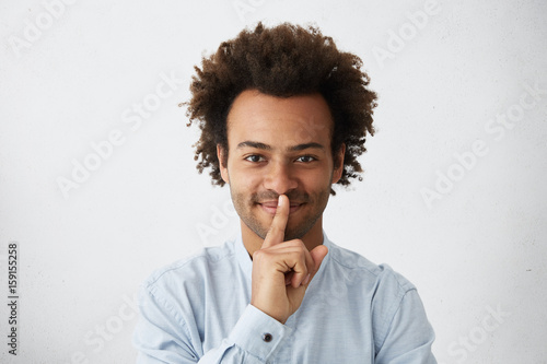 Happy dark-skinned guy with African hairstyle wearing elegant white shirt showing silence sign asking to be quiet. Indoor portrait of mixed race man holding his finger on lips having pleasant look