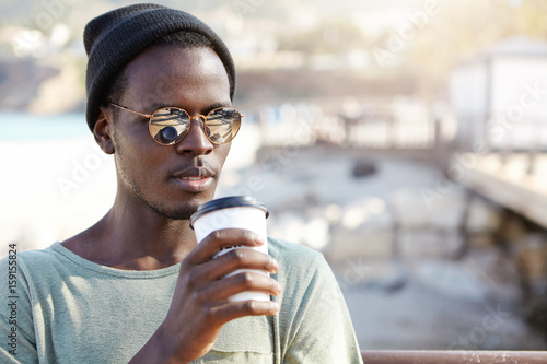 Handsome man in trendy sunglasses standing in open air holding cup of coffee enjoying its taste. Fashionable tourist in black hat standing in big city drinking hot tea having rest after long walk