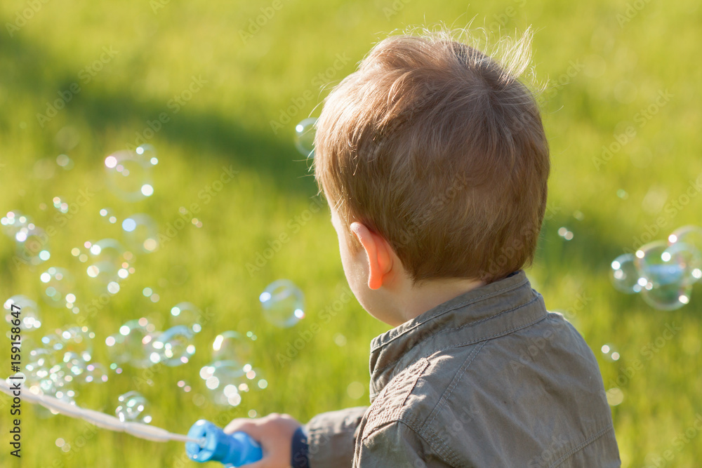 A little boy blowing soap bubbles in summer park. Background of grass outdoor.