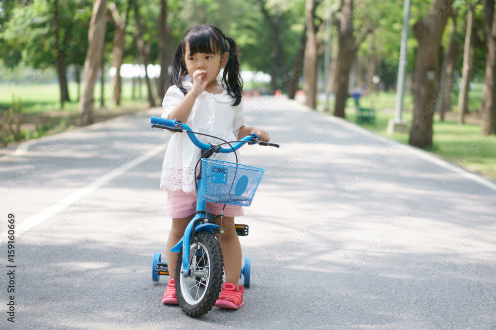 Asian children cute or kid girl cycling on blue bike or bicycle and nail biting on the road at public park with green tree for enjoying and exercise on summer holiday Asian children cute or kid girl cycling on blue bike or bicycle and nail biting on the road at public park with green tree for enjoying and exercise on summer holiday