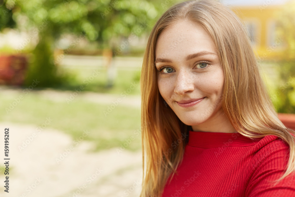 Pretty young blonde girl with green eyes, thin lips, dimples on cheeks and freckles on face wearing red clothes looking happily into camera while sitting against green background admiring nature Pretty young blonde girl with green eyes, thin lips, dimples on cheeks and freckles on face wearing red clothes looking happily into camera while sitting against green background admiring nature