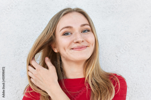 Pleased woman with blonde hair, luminous eyes and gentle smile having dimples on cheeks touching her beautiful hair with hand dressed in red clothes having good mood while posing against white wall photo