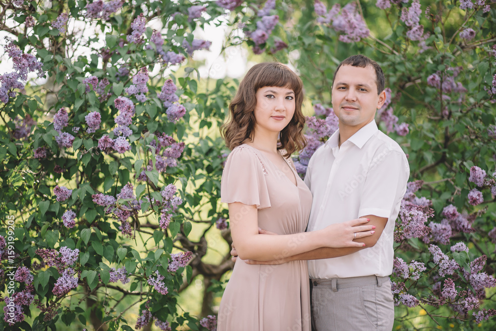 beautiful young couple in bright clothes, dress, shirt, couple, watching with a smile, hug, near the tree lilac in nature. in the Park, summer, greens, day, horizontal