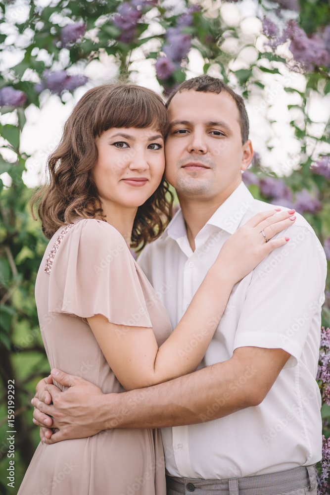 Fototapeta premium beautiful young couple, the girl in beige light dress, the guy in the white shirt, cuddling, watching with a smile near the tree, greens, summer, lilac, close-up, nature