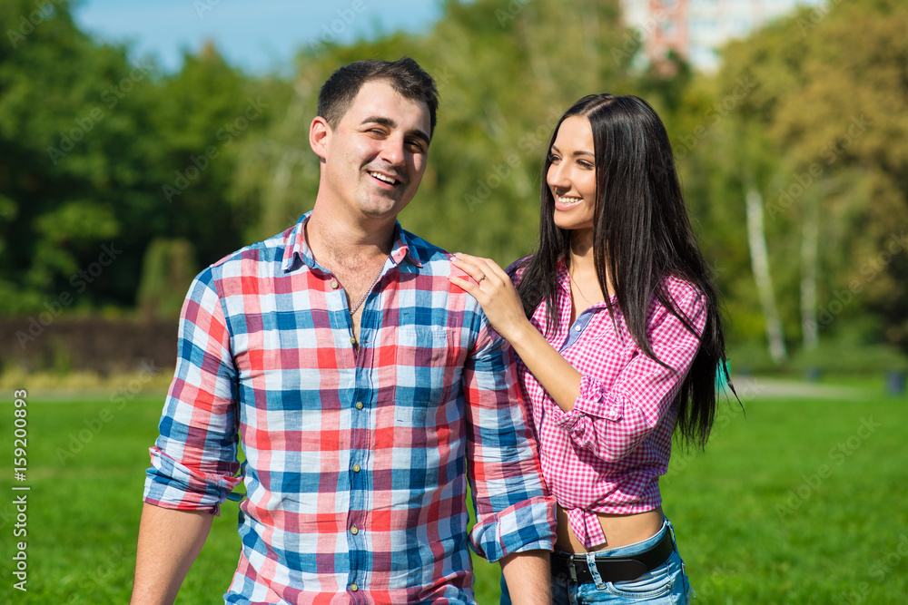 Young beautiful loving couple in checked shirts and jeans standing on the green lawn and laughing. A girlfriend is holding her hand on her boyfriend’s shoulder. Outdoor shot Young beautiful loving couple in checked shirts and jeans standing on the green lawn and laughing. A girlfriend is holding her hand on her boyfriend’s shoulder. Outdoor shot