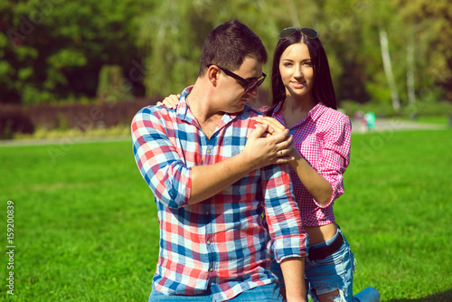Young beautiful loving couple in checked shirts, jeans and sunglasses sitting on the green lawn. A boyfriend holding his girlfriend’s hand on his shoulder and looking at it. Outdoor shot