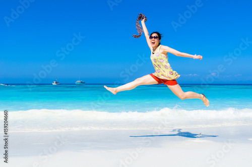 Happy and Cheerful beautiful Asian woman enjoy jumping on a white sand beach holding the blowing scarf with blue sky and blue sea as a background