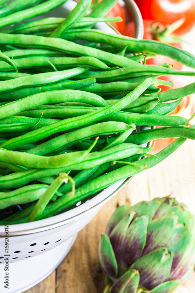 Bunch of fresh raw green beans in white metal colander on plank wood kitchen table, tomatoes, artichokes, preparing dinner, healthy diet, clean eating