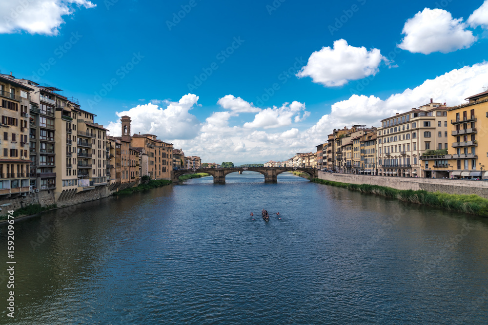 Horizontal composition the River Arno in Florence with puffy clouds and rowers on the water.
