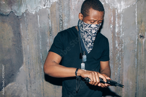 Black gang member checks his weapon. Gangster man with gun in hand on dark gray background. Outlaw, ghetto, murderer, armed attack concept photo