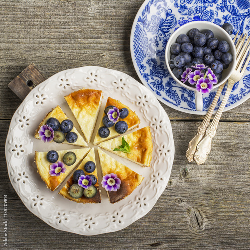 Homemade fresh cheesecake with blueberries and edible flowers on a white ceramic plate, on a simple wooden background. From the top view. The concept is helpful comfortable food