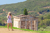 Travel woman photographer in greek dress takes shot of Archaeological Site of Ancient Messene, Peloponnese, Greece. Seductive female photographing a Greek Temple. The Mausoleum on blurred backgroung.