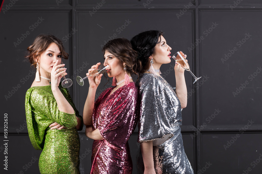 three beautiful girls girlfriend brunette in sparkly holiday dresses stand near a grey wall with wine glasses of champagne, the bachelorette party