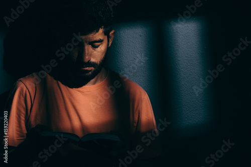 Prisoner man in dark cell reading a book or bible