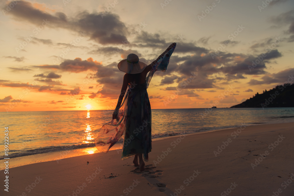 Back view of lonely girl with summer dress and hat is walking along coastline. Silhouette young woman on the beach at sunset. 