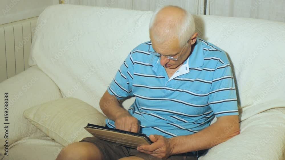 Elderly man uses tablet. Aged gray-haired male with eyeglasses types message on tablet PC concentrating on screen sitting on couch at home. Elderly man uses tablet. Aged gray-haired male with eyeglasses types message on tablet PC concentrating on screen sitting on couch at home.