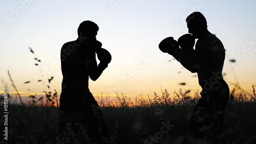Boxing training outdoors. Opponents in boxing gloves silhouette sparring in field at sunset. Fighter kicking.