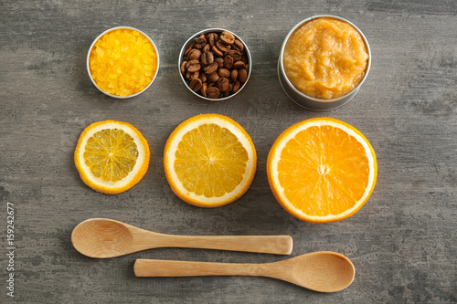 Jar with scrub, wooden spoons, sea salt, coffee grains and orange slices on grey wooden background