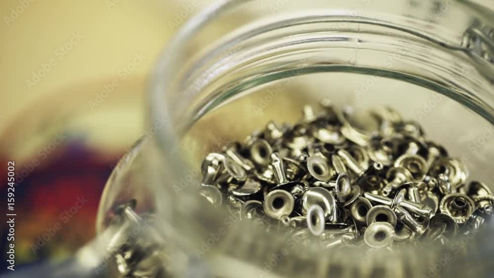 Glass open jar full of metal cloth rivets on desk in work shop, close up slow motion shot Glass open jar full of metal cloth rivets on desk in work shop, close up slow motion shot