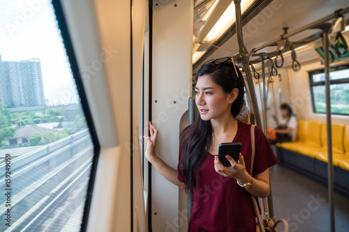 Asian woman passenger with casual suit using smart mobile phone in the BTS Skytrain rails or MRT subway for travel in the big city, lifestyle and transportation concept