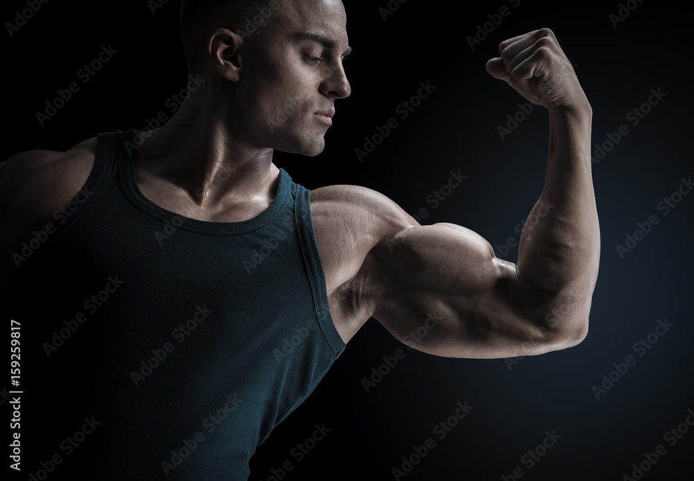 Young confindent muscular bodybuilder guy standing on black background and posing biceps muscle Classic bodybuilding