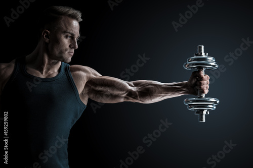 Male bodybuilder workout pumping up muscles holding dumbbell stretching out his hand. Strong athlete with perfect deltoid muscles, shoulders, biceps, triceps and chest.