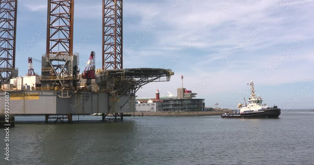 Jack up rig Atlantic Rotterdam, Three-Legged Self Elevating Unit drilling platform, berthed in Haringhaven. Tug Triton sailing by. IJMUIDEN, THE NETHERLANDS - MAY 2017 Jack up rig Atlantic Rotterdam, Three-Legged Self Elevating Unit drilling platform, berthed in Haringhaven. Tug Triton sailing by. IJMUIDEN, THE NETHERLANDS - MAY 2017